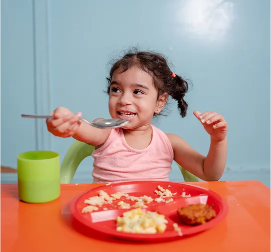 Niña pequeña sonriente comiendo con cuchara frente a un plato y un vaso sobre una mesa naranja.