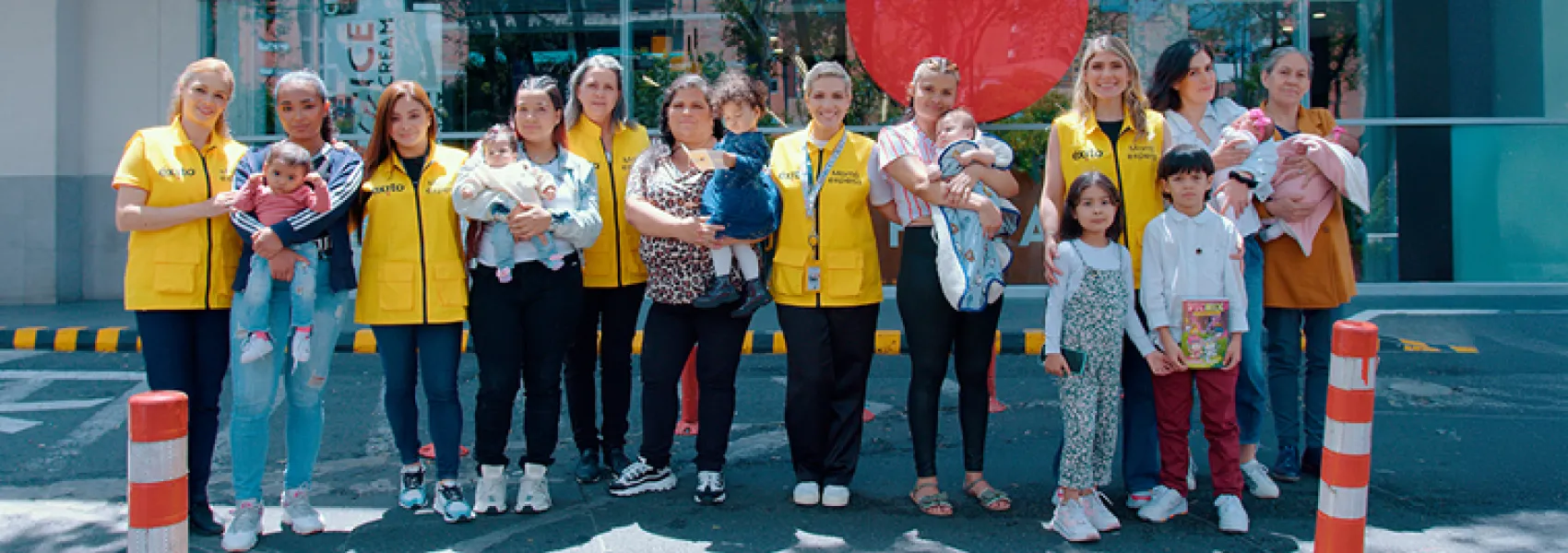 Un grupo de mujeres con chalecos amarillos sosteniendo bebés, junto a otras madres y varios niños, posando frente a un edificio en una calle.